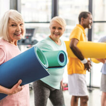 selective focus of senior sportswoman holding fitness mat and her friends standing behind at gym
